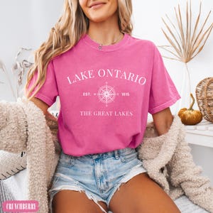 May include: A bright pink short-sleeve t-shirt featuring white text that reads LAKE ONTARIO, a compass graphic with EST. 1615, and the text THE GREAT LAKES. The shirt has a crew neckline and a relaxed fit, styled with light blue denim shorts.