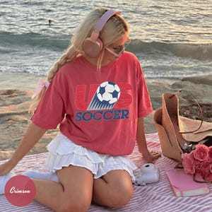 May include: A red t-shirt with the text "US SOCCER" and a soccer ball graphic. The shirt is paired with a white skirt. Accessories include pink headphones, a straw tote bag, and a pink notebook.
