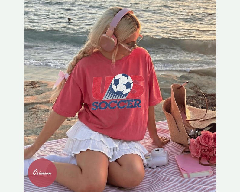 May include: A red t-shirt with the text "US SOCCER" and a soccer ball graphic. The shirt is paired with a white skirt. Accessories include pink headphones, a straw tote bag, and a pink notebook.