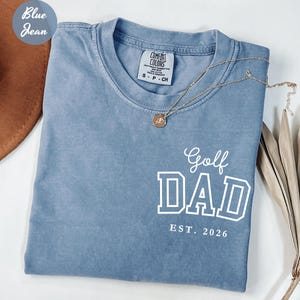May include: A blue jean-coloured t-shirt with the words "Golf DAD EST. 2026" in white block letters. The shirt has a crew neck and short sleeves. A gold necklace with a pendant is draped across the shirt.