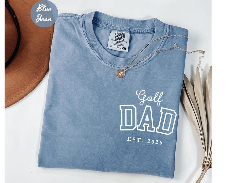 May include: A blue jean-coloured t-shirt with the words "Golf DAD EST. 2026" in white block letters. The shirt has a crew neck and short sleeves. A gold necklace with a pendant is draped across the shirt.