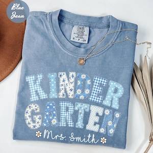 May include: A blue jean colored t-shirt featuring the word KINDERGARTEN in large, patchwork-style letters with various blue and white floral and gingham patterns. Below the text, the name Mrs. Smith is printed in white script with small daisy icons.