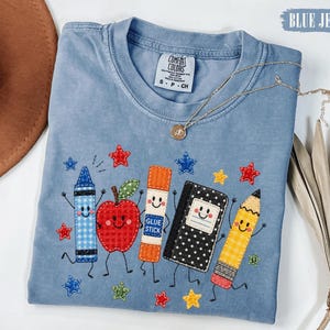 May include: A blue crew neck t-shirt featuring an embroidered graphic of a blue crayon, a red apple, an orange glue stick, a black and white polka dot notebook, and a yellow pencil, all with smiling faces, surrounded by small colourful stars.