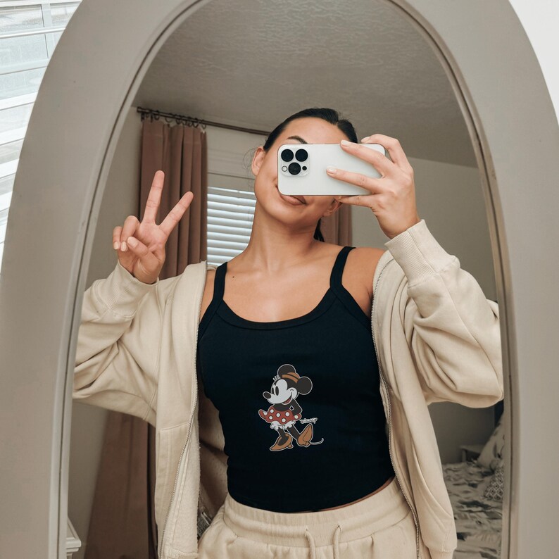 May include: A black ribbed tank top featuring a graphic of Minnie Mouse wearing a red polka dot dress and brown shoes, layered under a beige zip-up hoodie. The tank top has a scoop neckline and thick straps, providing a casual look.