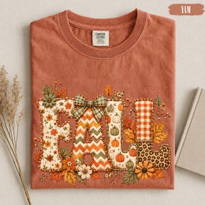 May include: A terracotta-coloured t-shirt featuring the word FALL in large, patterned letters. The F has a floral print, the A has a chevron pattern with a plaid bow, the L has a pumpkin print, and the second L has a plaid and leopard print.