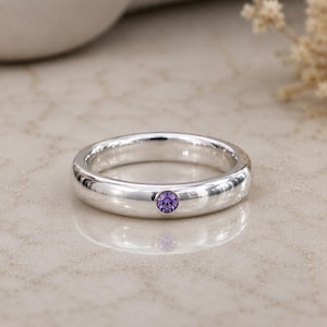 Peut inclure: Une bague anneau en argent poli ornée d'une unique petite pierre précieuse ronde de couleur violette, sertie à fleur dans le métal. La bague présente une finition lisse et réfléchissante ainsi qu'un design circulaire simple et uniforme.