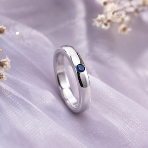 May include: A polished silver-toned band ring featuring a single, small, round-cut blue gemstone set flush into the metal surface. The ring has a smooth, rounded profile and a reflective finish, showcasing a clean and simple design aesthetic.