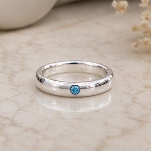 May include: A polished silver band ring featuring a single, small, round, bright blue gemstone set flush into the center of the metal surface. The ring has a smooth, rounded profile and a reflective finish that catches the light.