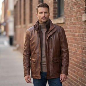 May include: A brown leather jacket with a high collar, a vertical zippered chest pocket, and two large flap pockets at the waist. The jacket is worn over a tan turtleneck sweater and blue denim jeans, featuring visible stitching details.