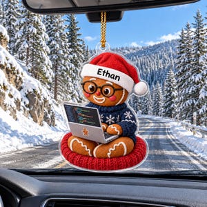 May include: A clear acrylic hanging ornament featuring a gingerbread person wearing a red Santa hat with the name Ethan printed on it, black glasses, and a blue snowflake sweater, sitting on a red cushion while using a laptop with code on screen.