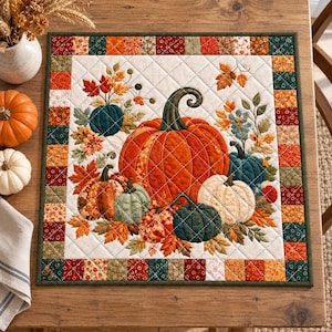 May include: A square quilted fabric mat featuring a central illustration of various pumpkins in orange, white, and teal, surrounded by autumn leaves. The border consists of a patchwork of small squares in shades of red, orange, green, and cream.