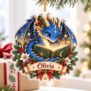 May include: A clear acrylic Christmas ornament featuring a blue dragon reading a glowing book. The design includes holly, red berries, white poinsettias, and a gold banner with the name Olivia printed in red. A red bow hangs below the banner.