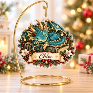 May include: A clear acrylic Christmas ornament hanging from a gold metal stand. The ornament features a sleeping teal dragon with gold accents resting on a dark green book. A banner at the bottom displays the name Chloe in red script.