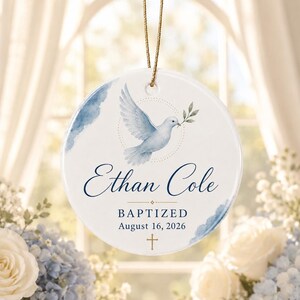 May include: A round white ceramic ornament featuring a watercolor illustration of a dove carrying an olive branch. The text reads Ethan Cole, BAPTIZED, and August 16, 2026, with a small gold cross at the bottom and a gold hanging string.