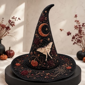 May include: A black quilted witch hat featuring intricate embroidery of a crescent moon, a large moth, pumpkins, roses, and spiderwebs. The design incorporates warm orange, cream, and deep red thread colors against the dark fabric base.