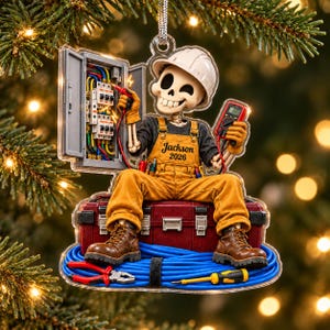 May include: A clear acrylic ornament featuring a skeleton wearing a white hard hat and yellow overalls with the text Jackson 2026. The skeleton holds a multimeter and electrical probes near an open circuit breaker panel, sitting on a red toolbox.