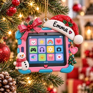 May include: A pink and blue tablet-shaped Christmas ornament featuring a screen with colorful app icons, a red bow, a snowman, and a Santa hat with the name Emma written on it. A small pink heart charm with a snowflake hangs from the side.
