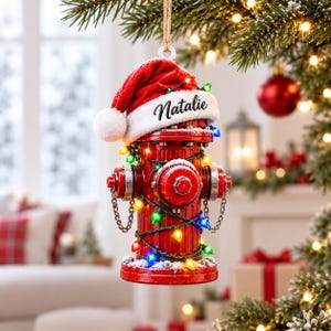 May include: A red fire hydrant Christmas ornament featuring a white-trimmed red Santa hat with the name Natalie printed on the brim. The hydrant is wrapped in a string of colorful miniature holiday lights and hangs from a brown twine loop.