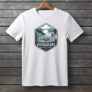 Peut inclure: Un t-shirt blanc à manches courtes avec un motif hexagonal central. Le dessin représente un castor brun nageant dans un lac entouré de pins verts et de montagnes, avec le texte VOYAGEURS NATIONAL PARK imprimé en dessous.