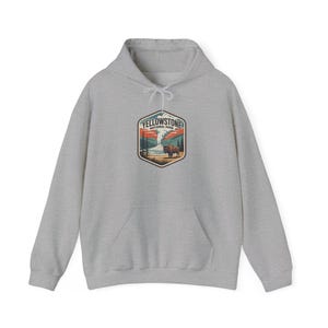 Puede incluir: Una sudadera con capucha gris claro con un gráfico de Yellowstone National Park en la parte delantera. El gráfico incluye un bisonte, un géiser y el texto "Yellowstone National Park". La sudadera tiene una capucha con cordón y un bolsillo delantero.