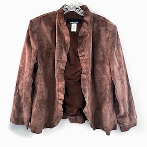 Puede incluir: Chaqueta de ante marrón sin cuello. La chaqueta tiene mangas largas y una parte delantera fluida y abierta. El forro interior es de un tono marrón más oscuro. La chaqueta cuelga sobre un fondo blanco.