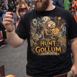 May include: Black t-shirt featuring a fantasy illustration with the text "THE HUNT FOR GOLLUM". The design includes Gollum, a warrior, and a glowing orb. The shirt is worn by a person holding a red die.