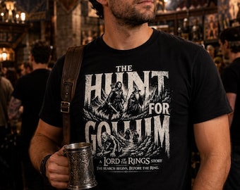May include: A black short-sleeved t-shirt featuring a white graphic print with the text THE HUNT FOR GOLLUM A LORD OF THE RINGS STORY THE SEARCH BEGINS. BEFORE THE RING. The design depicts characters in a mountainous landscape.
