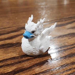 以下が含まれることがあります： 青い嘴と詳細な翼を持つ白いドラゴンの置物。 ドラゴンはテクスチャ加工された表面を持ち、座った姿勢です。 この置物はユニークな装飾品です。