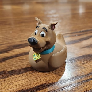 Op de afbeelding: Een bruine 3D-geprinte Scooby-Doo badeend met een zwarte neus, witte ogen en een blauwe kraag met een gele tag. De eend staat op een houten oppervlak.