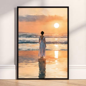May include: A framed wall art print depicting a person standing on a wet sandy beach at sunset. The person wears a long, flowing white dress. The scene features a bright orange sky, a glowing sun, and ocean waves reflecting the warm light.