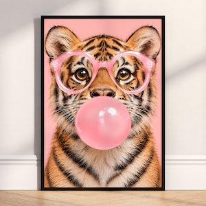 May include: A framed wall art print featuring a tiger cub with orange and black stripes, wearing round pink glasses and blowing a large pink bubblegum bubble. The background is a solid light pink colour, and the frame is black.