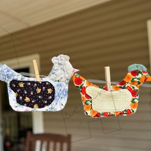 May include: Three handmade chicken-shaped fabric items hanging on a clothesline. The chickens have different patterned fabrics, including floral, purple, and fruit designs. Each is held by a wooden clothespin, showcasing a rustic, country aesthetic.