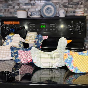 May include: Four fabric chicken-shaped kitchen items arranged in a row. Each features a unique pattern, including beige stripes, navy blue floral, light green plaid, and a vibrant orange and blue floral print with a yellow gingham wing detail.