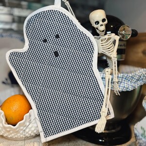 以下が含まれることがあります： 黒と白のギンガムチェック柄のゴースト型オーブンミット。目と口に見立てた3つの小さな黒い円形のアクセントが付いています。ミットはキルティング加工が施され、縁には白いトリミングがあり、上部には吊り下げ用の小さな布製ループが付いています。
