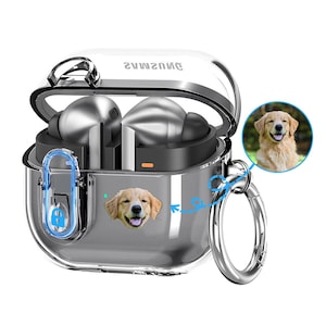 May include: Clear protective case for wireless earbuds with a silver carabiner clip. The case features a smiling golden retriever graphic and the word "samsung" printed on the top. The earbuds are silver.