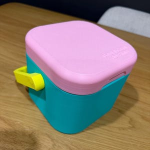 May include: A small, square container with a pink lid and a teal base. The container has a yellow handle on the side. The lid has the text "Creative Design Studio" printed on it. The container is on a wooden surface.