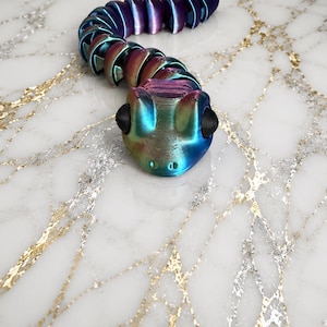 May include: A 3D-printed articulated snake toy in iridescent colors of blue, purple, and bronze. The snake's head has black eye details. The toy is positioned on a white marble surface with gold veining.