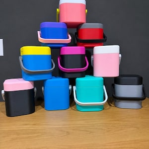 May include: A selection of small, square coolers in various colours, with handles. The coolers are stacked on a wooden surface against a dark grey background. Colours include pink, blue, yellow, black, red, and teal. The coolers have white handles.