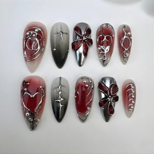 Peut inclure: Un ensemble de dix faux ongles avec divers motifs. Les ongles présentent un dégradé de couleurs, notamment du rouge, de l'argent et du gris. Certains ongles comportent des ornements argentés, notamment des cœurs, des nœuds et des formes géométriques.