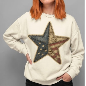 May include: Cream-coloured sweatshirt with a large star design. The star is divided into sections with a blue field of stars, red and white stripes, and decorative stitching. The design has a vintage, patriotic aesthetic.