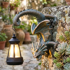 Könnte beinhalten: Eine bronzefarbene Drachenskulptur hält eine schwarze Laterne an einer Steinmauer. Der Drache hat detaillierte Flügel und einen eingerollten Schwanz. Die Laterne hat ein warmes, leuchtendes Licht. Der Hintergrund zeigt einen Garten.