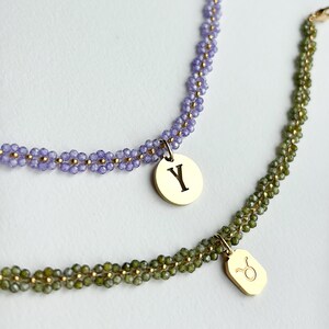 Puede incluir: Dos collares de cuentas. Uno es lavanda con un colgante dorado con la letra "Y". El otro es verde oliva con un colgante dorado con un símbolo del zodiaco Tauro. Ambos collares tienen detalles dorados.