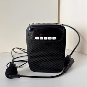 Puede incluir: Amplificador de voz portátil negro con altavoz y micrófono incorporados. El dispositivo tiene forma rectangular con una parte frontal perforada y un panel de control con botones. Un cable negro conecta el micrófono al amplificador.