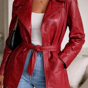 May include: A red leather jacket with a belted waist and notched lapels. The jacket has a classic trench coat design, with a double-breasted front and pockets. The model is wearing a white top and blue jeans. A black purse with a gold chain is on the left shoulder.