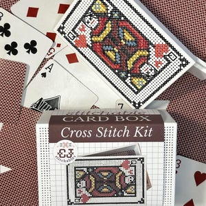 May include: A cross stitch kit for a card box featuring a playing card design. The kit includes a white box with a grid pattern, a label reading "Stitchable CARD BOX Cross Stitch Kit" with a logo, and a stitched image of a king playing card.