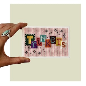 May include: A rectangular cross-stitch embroidery piece featuring the word Trinkets in colorful block letters. The background is a light pink and white vertical striped fabric. Small black cross-stitch star accents are scattered around the text.