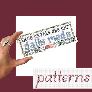 May include: A rectangular cross-stitch sampler featuring the text "Give us this day our daily meds" in dark blue and black thread, accented with a small red and white pill icon and green floral border motifs, presented on a white background.