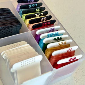 May include: A translucent plastic storage container divided into compartments, holding organized embroidery floss on plastic bobbins. The bobbins are black or white, each labeled with numerical identifiers like 210, 597, 15, 727, 20, 304, 33, 3846, 486, 729, 21, and 816.