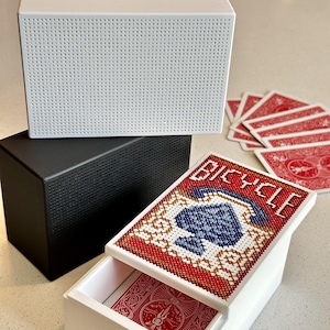 May include: A white rectangular box with a perforated texture, a black rectangular box with a perforated texture, and a white sliding box featuring a cross-stitch design of a blue spade and the word BICYCLE, containing a deck of red playing cards.