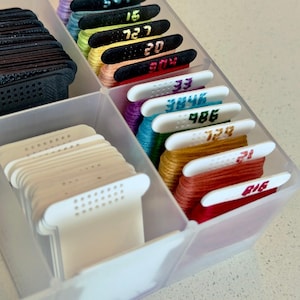 May include: A clear plastic storage container holding multiple embroidery floss bobbins. The bobbins are organized in rows, featuring various colors of thread wrapped around white or black plastic cards, each labeled with printed numbers like 15, 727, 20, 304, 33, 3846, 486, 728, 12, and 816.